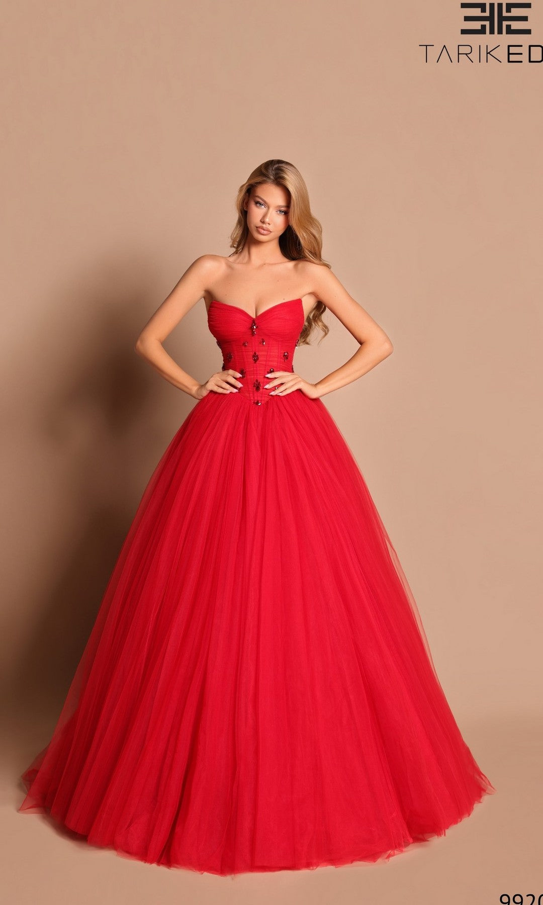 A woman poses against a plain beige background in the Red Corset Ball Gown Tarik Ediz 99205, featuring a fitted bodice and full tulle skirt.